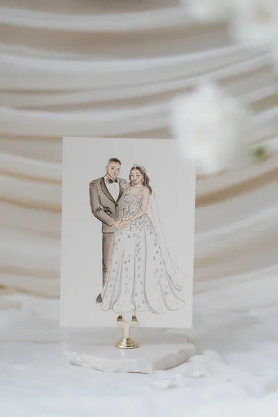 Watercolour and ink illustration of a bride and groom on a card held in a gold stand, by Adore by Lissy