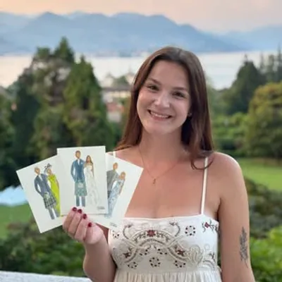 Artist Charlotte Beddoes holding three watercolour wedding guest illustrations by a lake view, by Charlotte Beddoes