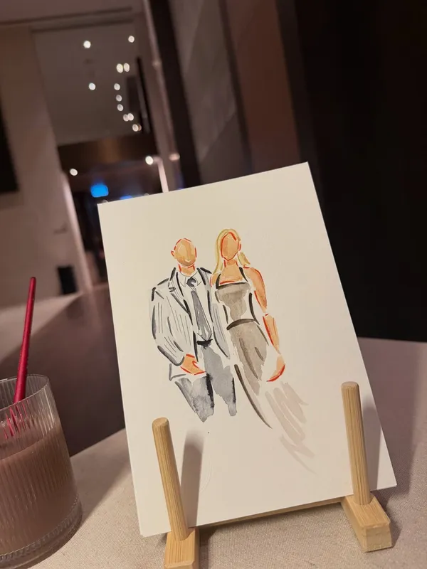 Faceless watercolour sketch of a wedding guest couple on a small wooden easel at a reception, by Watercolour Guest Portraits