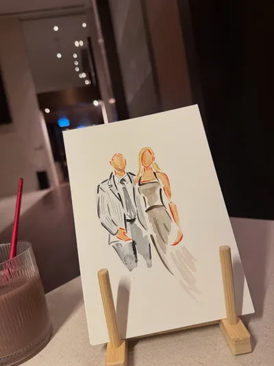 Faceless watercolour sketch of a wedding guest couple on a small wooden easel at a reception, by Watercolour Guest Portraits