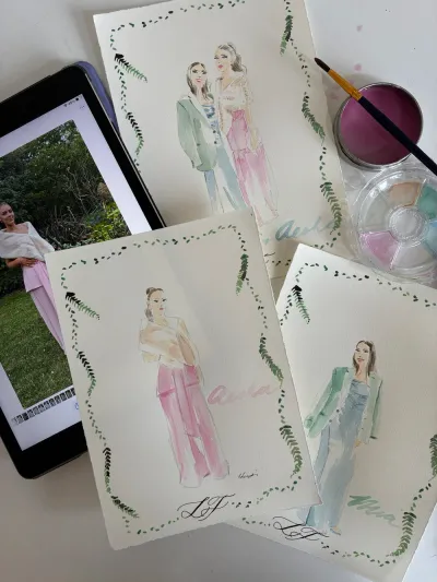 Watercolour fashion sketches of wedding guests on fern-bordered cards beside a paint palette and brush by Studio Heidi